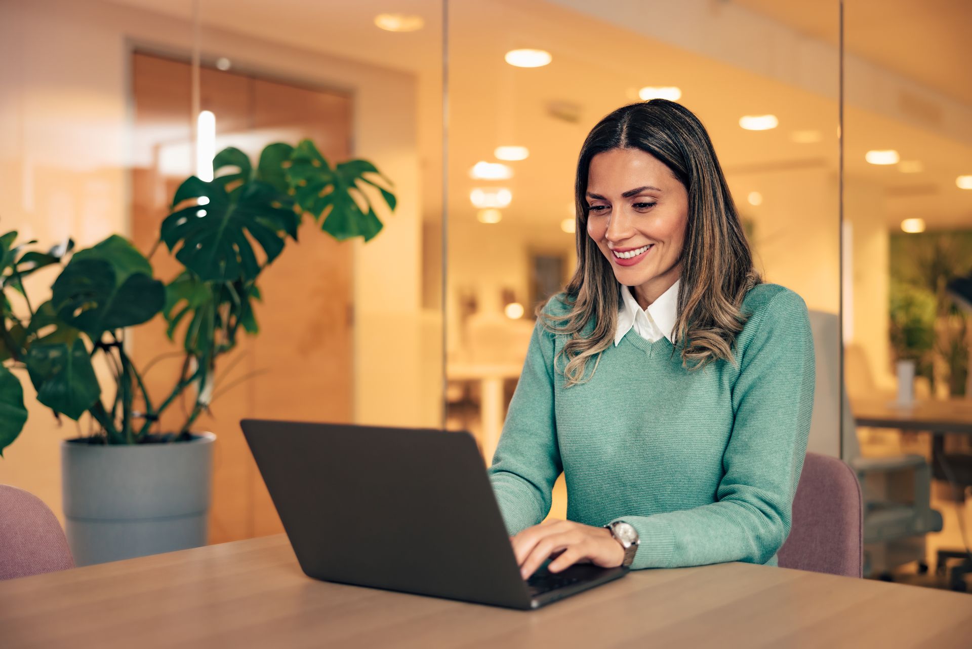 Woman smiling and typing on a laptop at a table in a warm office setting