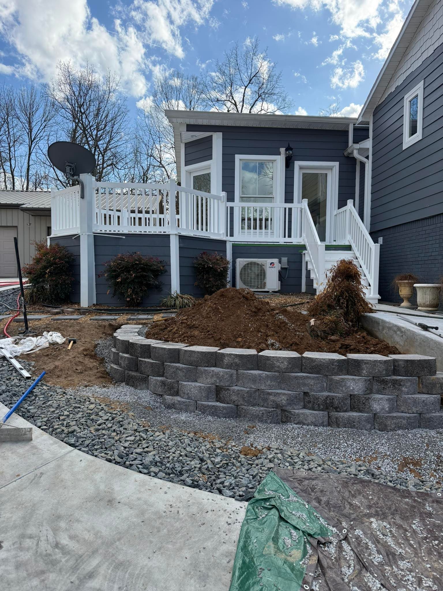 A dark gray house with a white deck, a stone retaining wall, and fresh dirt landscaping on a sunny day.