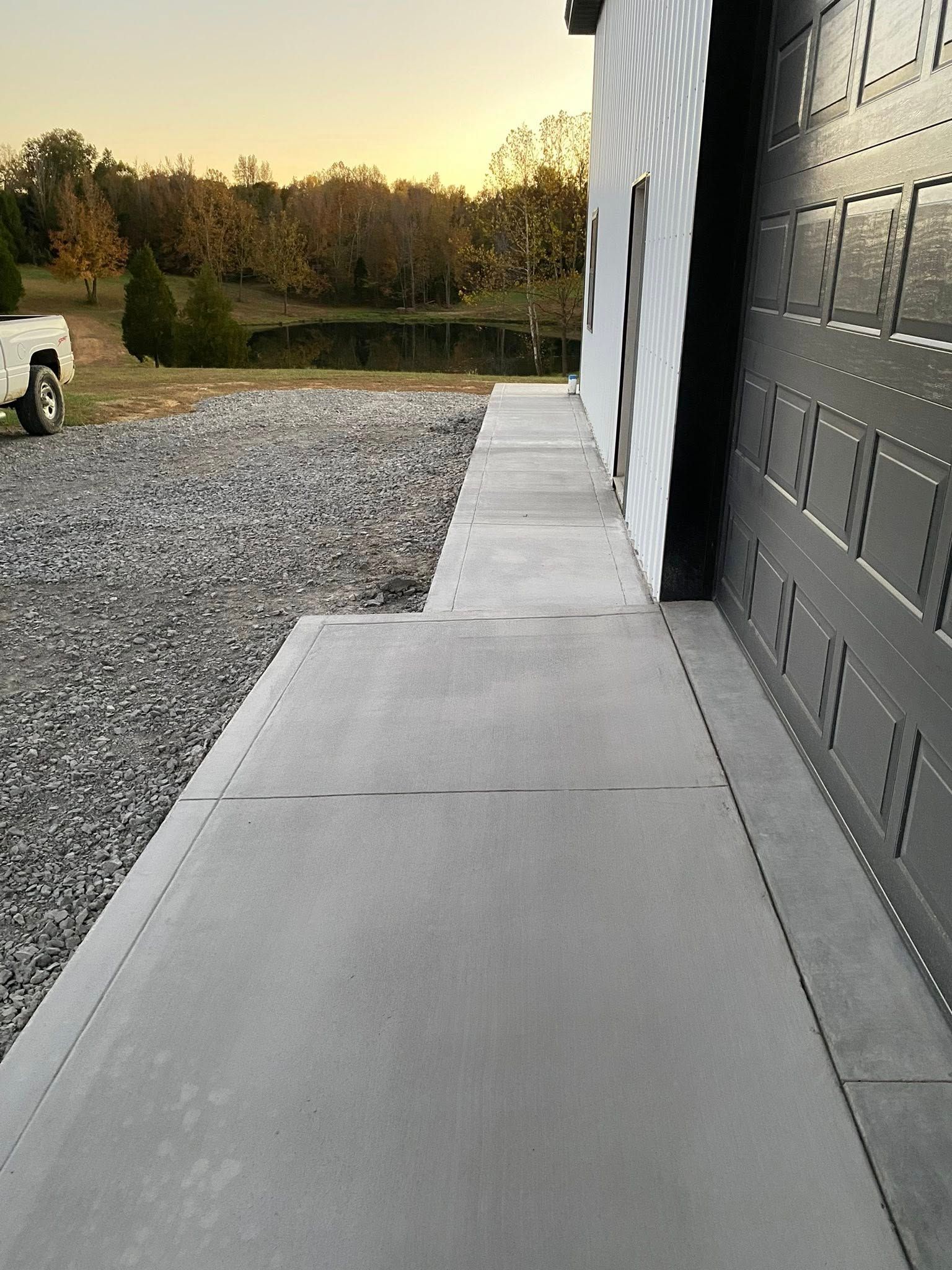 A concrete walkway runs along the side of a building with a gravel parking area and a pond in the background at sunset.