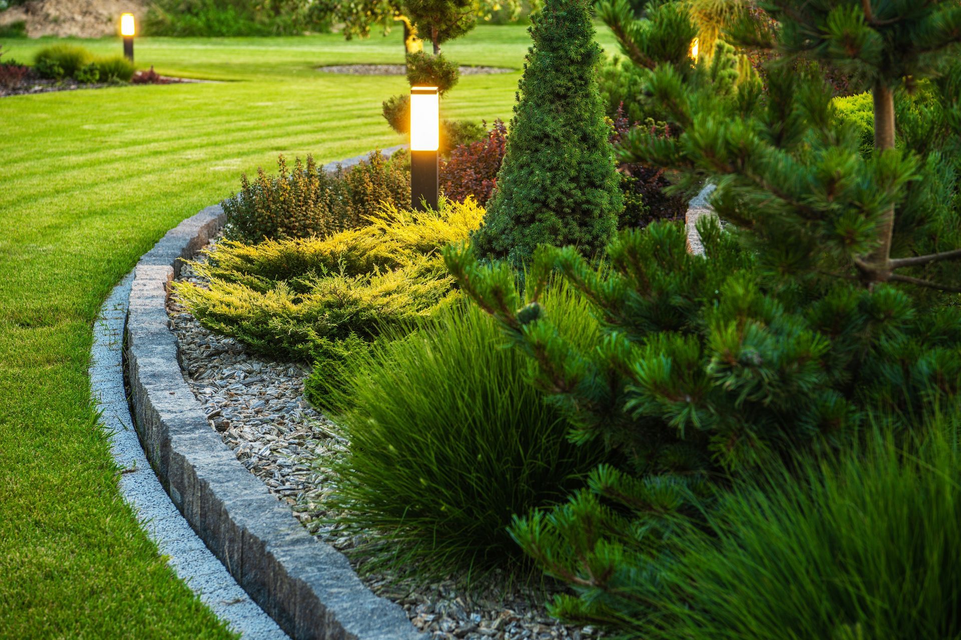 A curved stone garden bed with evergreen shrubs and illuminated path lights borders a manicured green lawn at twilight.
