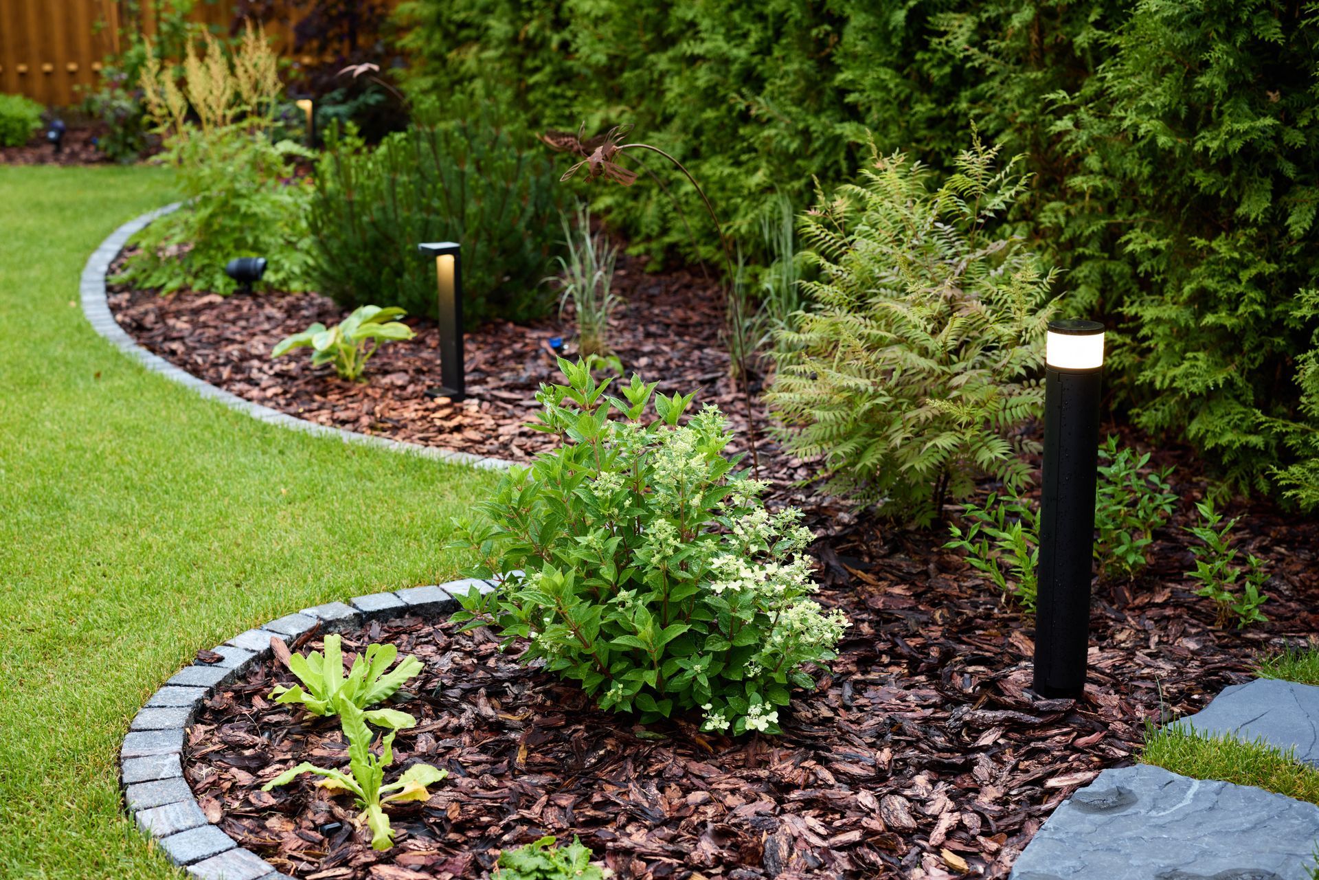 A curved garden bed featuring mulch, small shrubs, and modern black bollard lights, bordered by gray stone blocks.