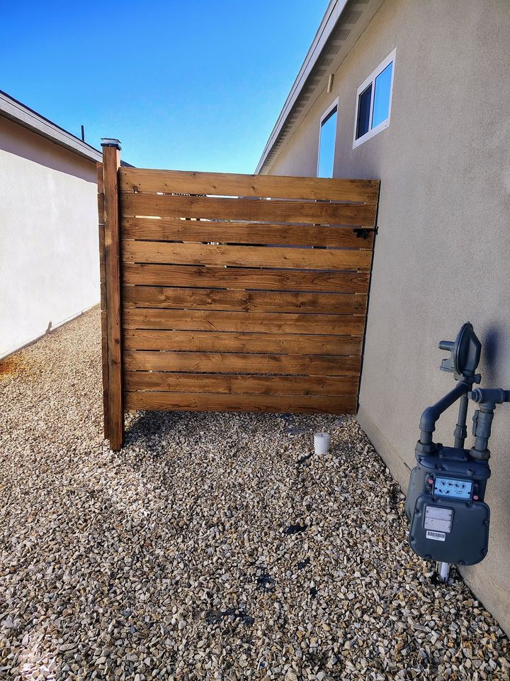 Wooden gate between stucco walls over gravel, with a blue water pump on the right.