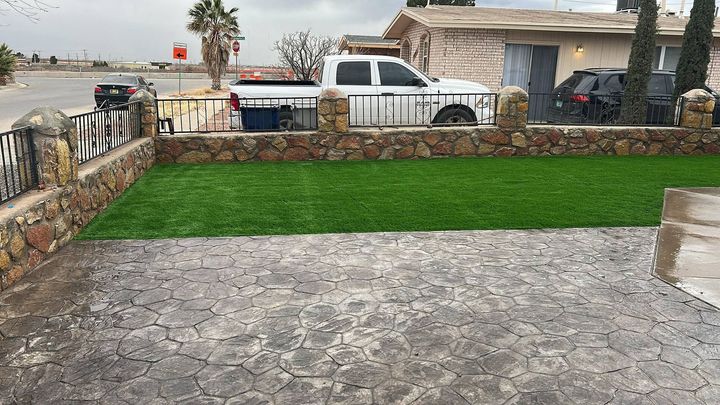 Stone patio and green lawn in a walled yard, with parked vehicles and a small building in the background