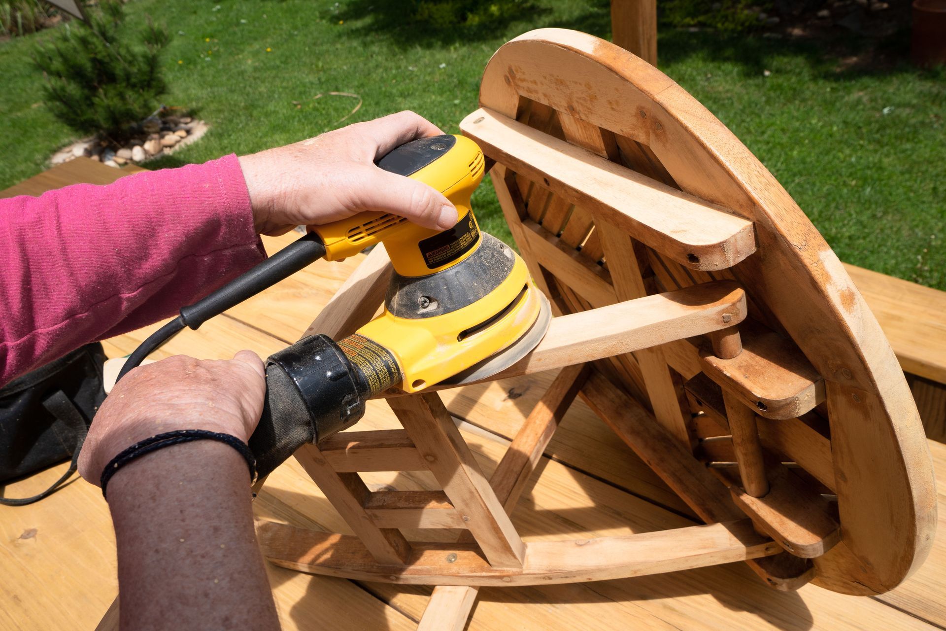 Hands sanding a wooden wheel assembly outdoors with a yellow electric sander.