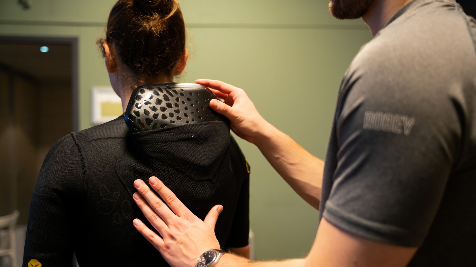 Een persoon in een zwart trainingspak, een andere persoon die de apparatuur op zijn rug aanpast, in een studio.