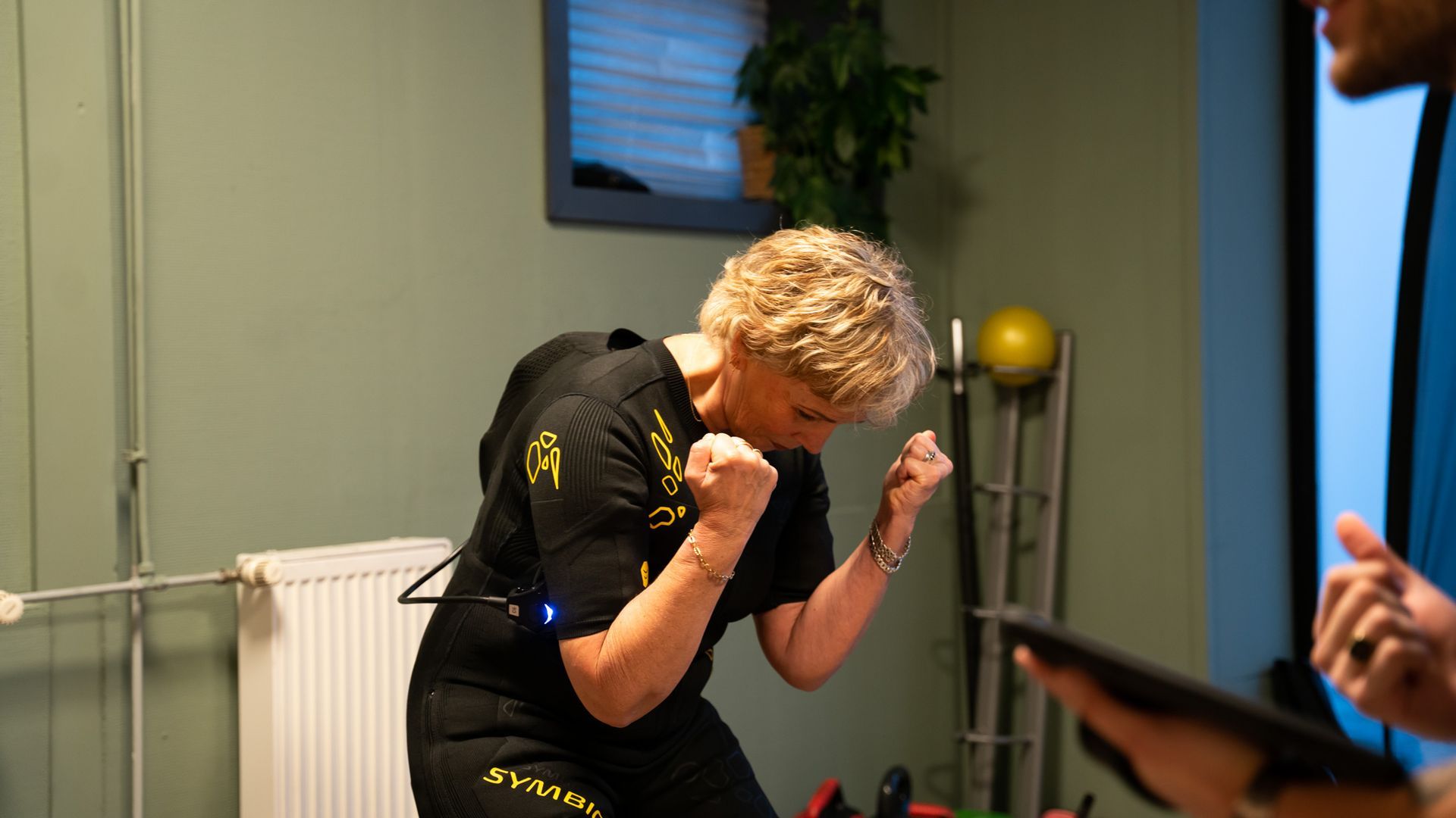 Vrouw in zwart pak met gebalde vuisten, mogelijk aan het sporten binnenshuis, groene muur op de achtergrond.