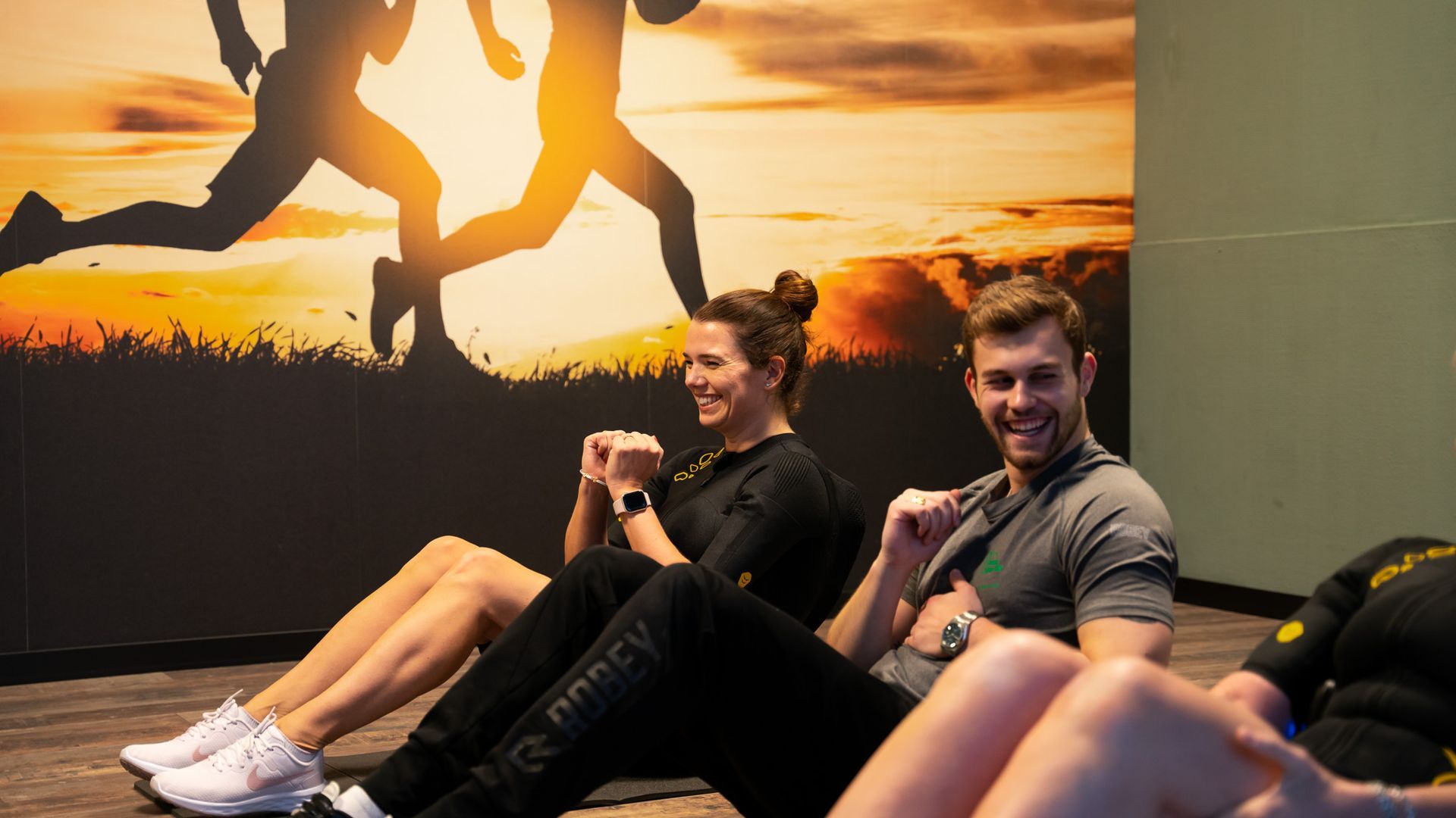 Twee mensen lachend terwijl ze sit-ups doen in een sportschool, met op de achtergrond een silhouet van een zonsondergang.