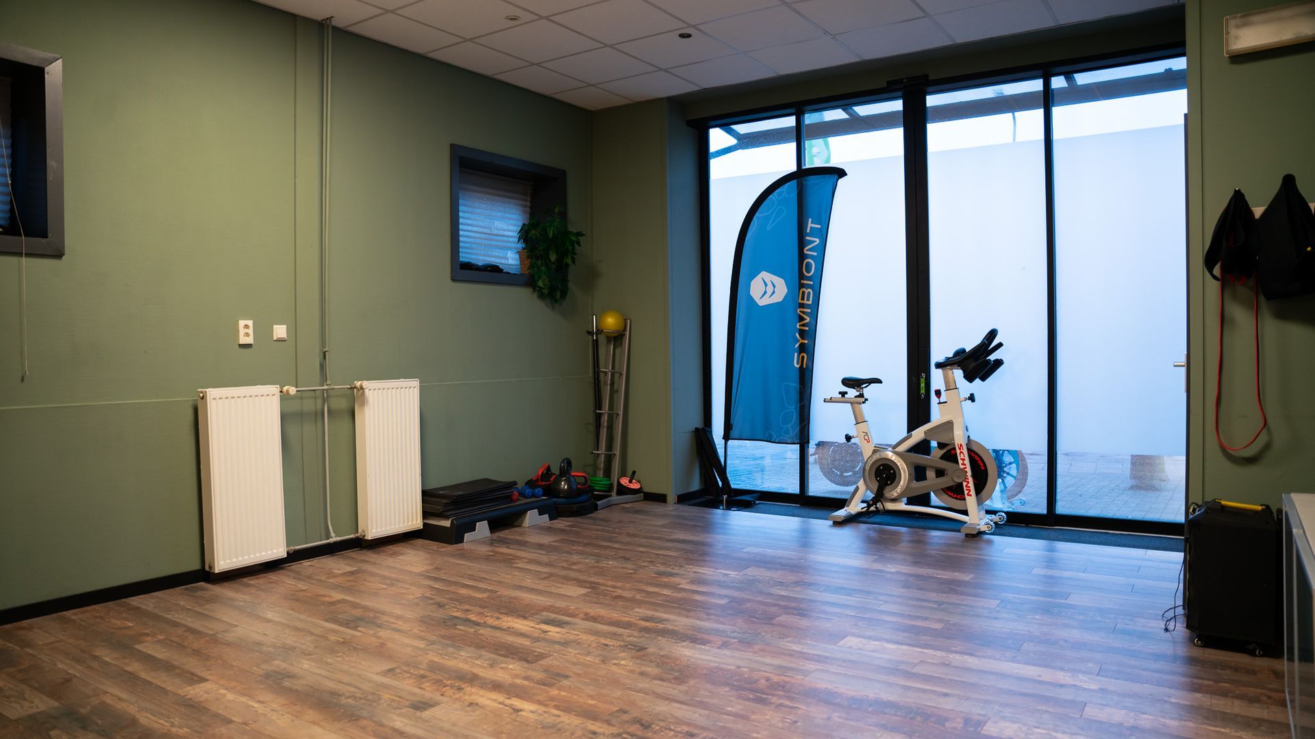 Interieur van een sportschool met een spinningfiets, gewichten en een spandoek naast een groot raam.