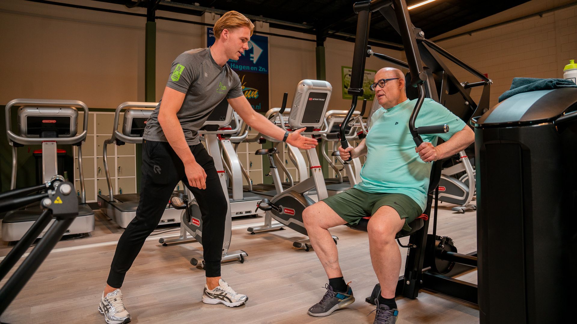 Een man staat naast een man die op een apparaat zit in een sportschool.