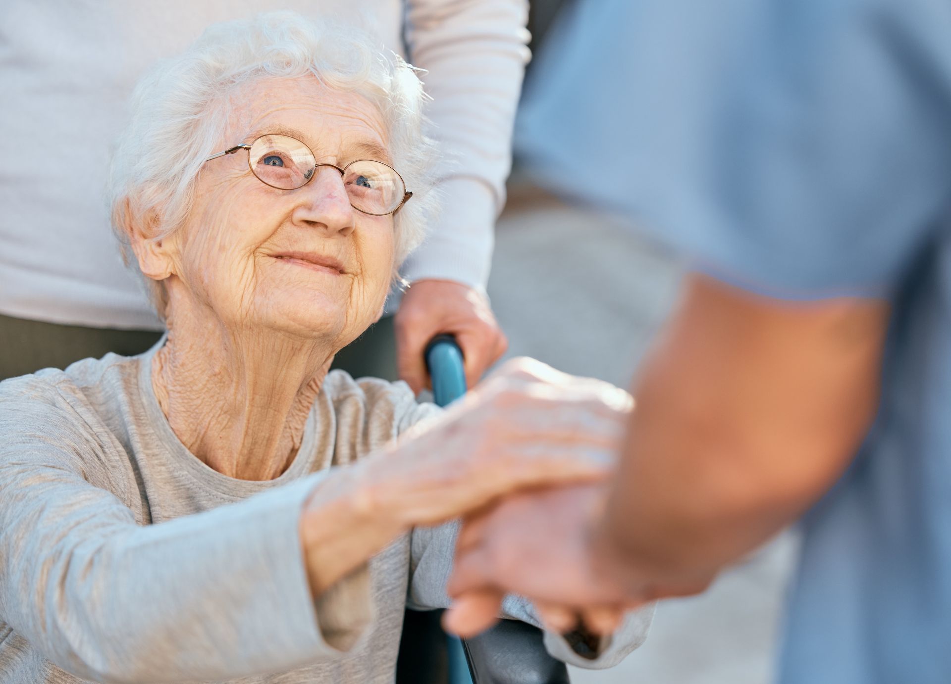 Elderly person in a wheelchair smiles as a person holds her hand; outdoors.