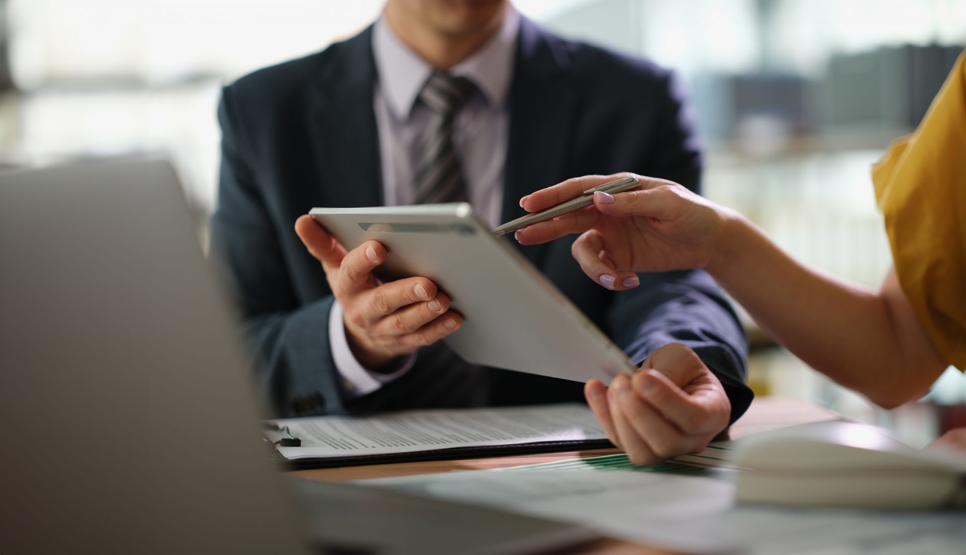 Man in suit and woman with yellow top reviewing a tablet together in an office.