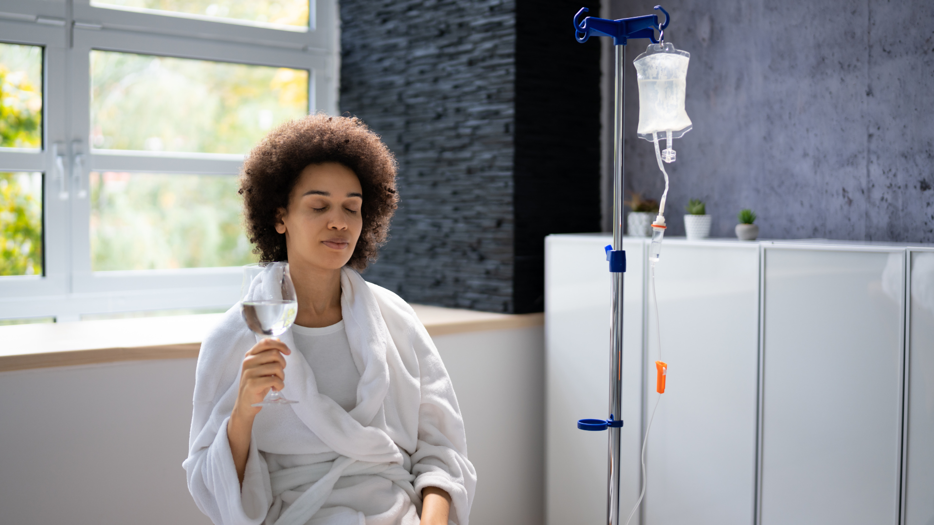 Woman in a white robe receives IV drip, holding a glass, in a well-lit room.