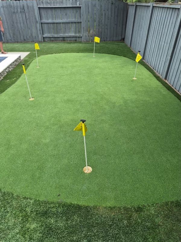 A backyard putting green with six yellow flags, surrounded by a dark wooden fence and grass.
