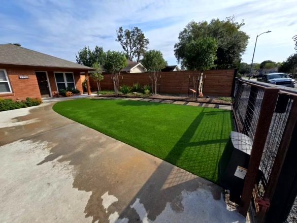 Front yard with green lawn, driveway, brick house, wooden fence, trees, and blue sky.