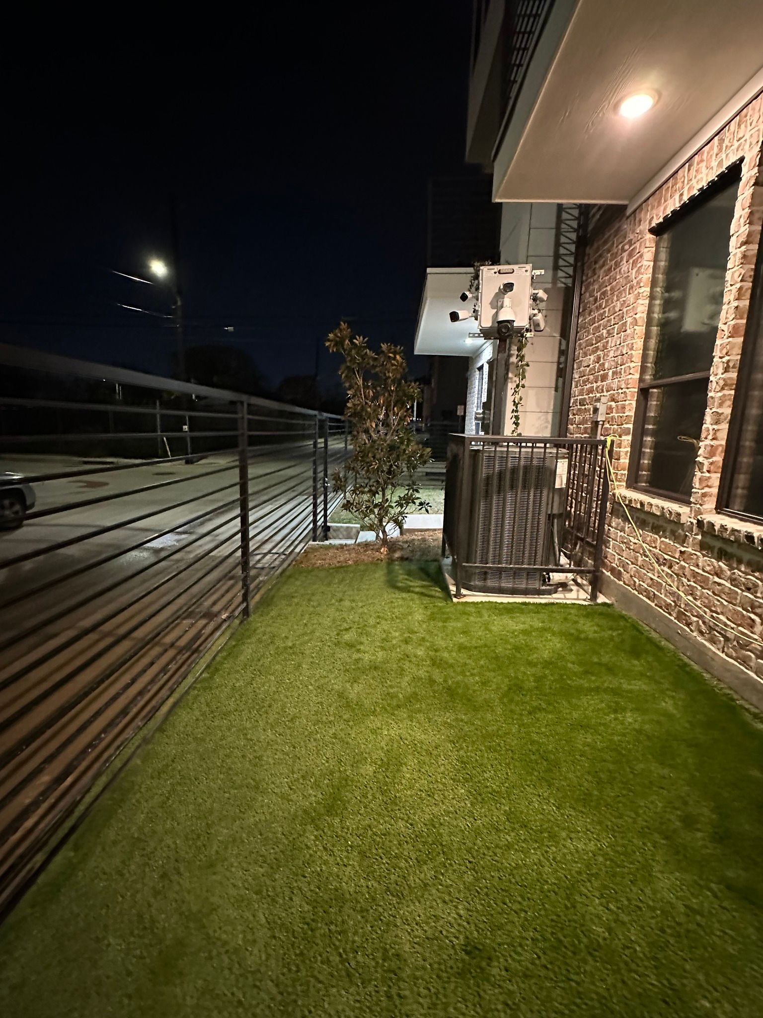 Exterior nighttime view of house with green lawn, brown brick wall, and a black metal fence.