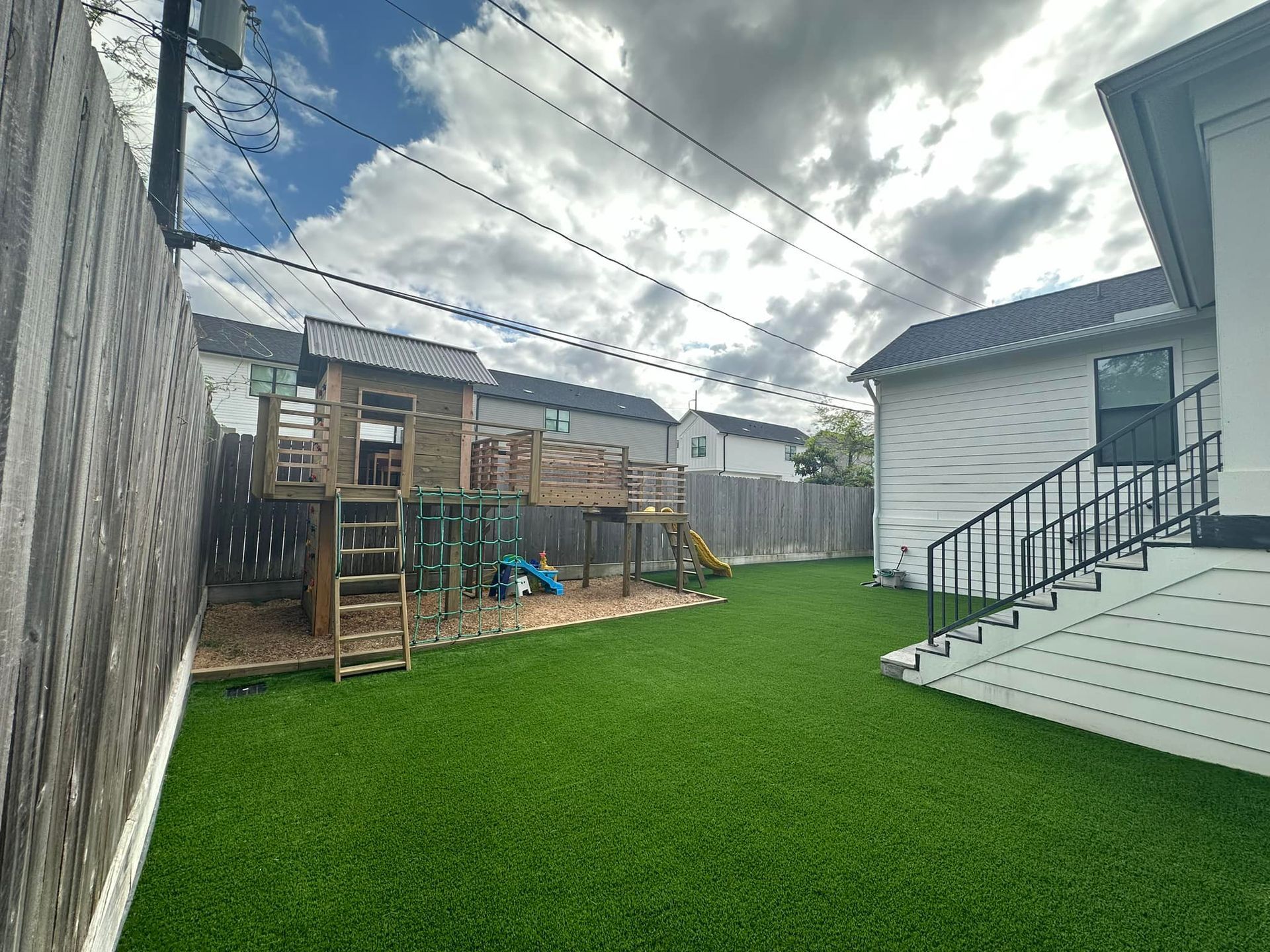 Backyard with play set, artificial grass, wooden fence, and white house with black stairs. Overcast sky.