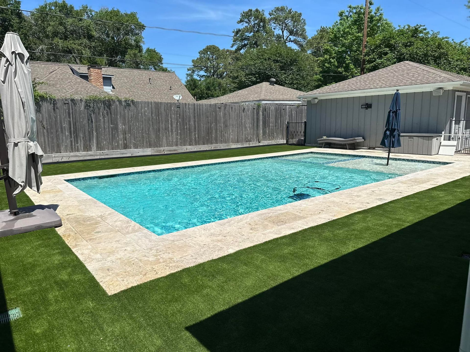 Rectangular turquoise pool surrounded by tan stone and green turf in a backyard setting.