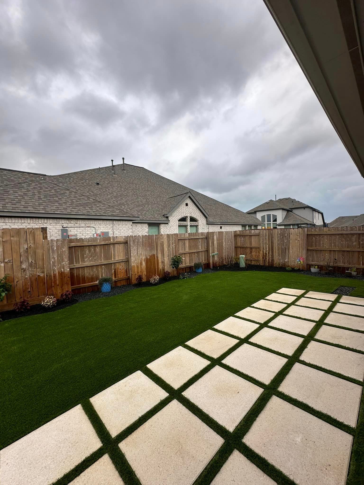 Backyard with green lawn, stone patio, wooden fence, and homes under cloudy sky.