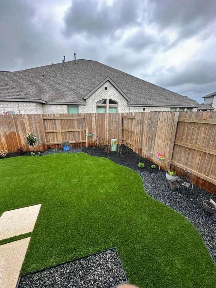 Backyard with green artificial turf, black rock border, wooden fence, and house under a cloudy sky.