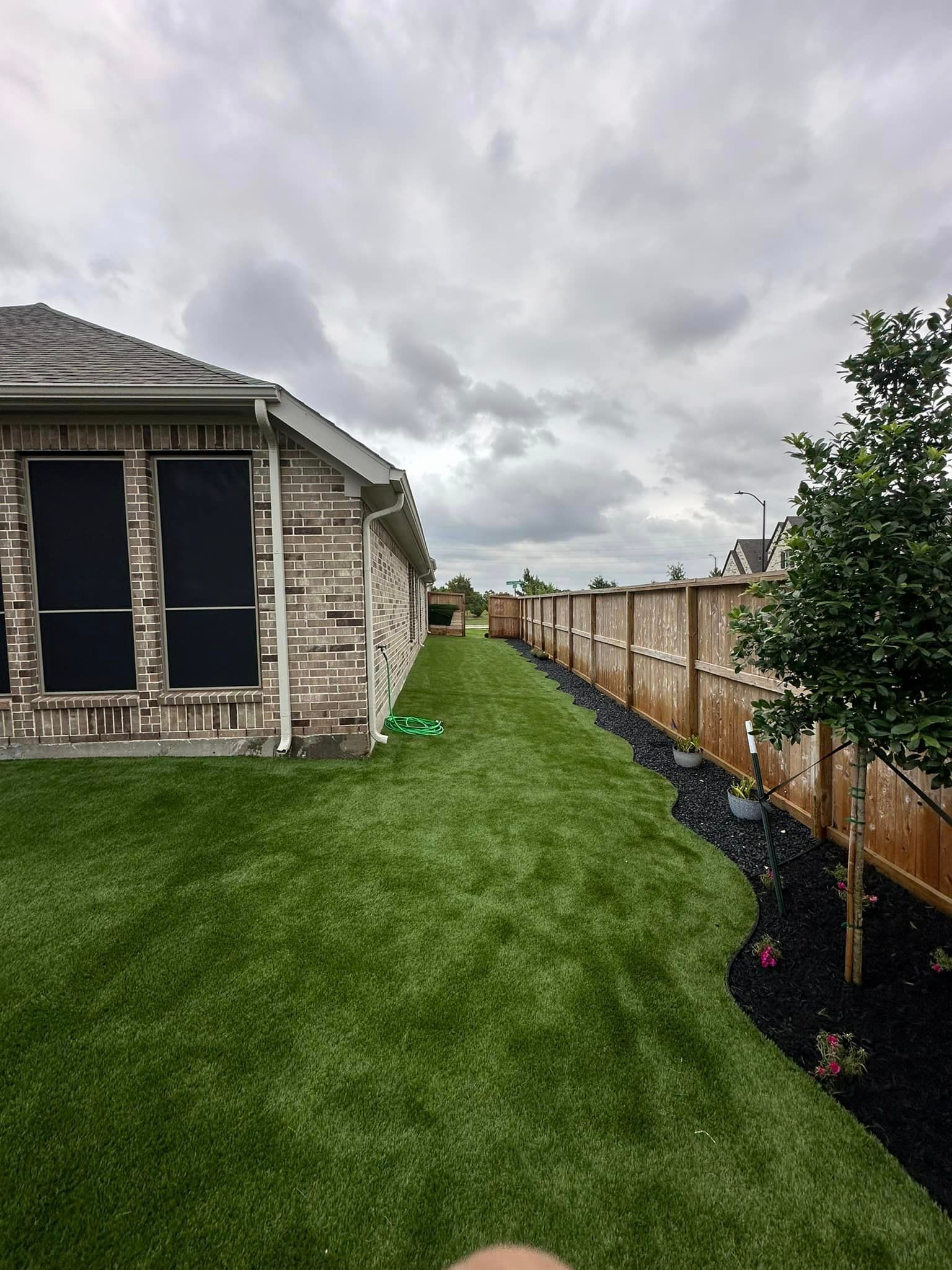 Backyard with green artificial turf, brown fence, brick house, and cloudy sky.