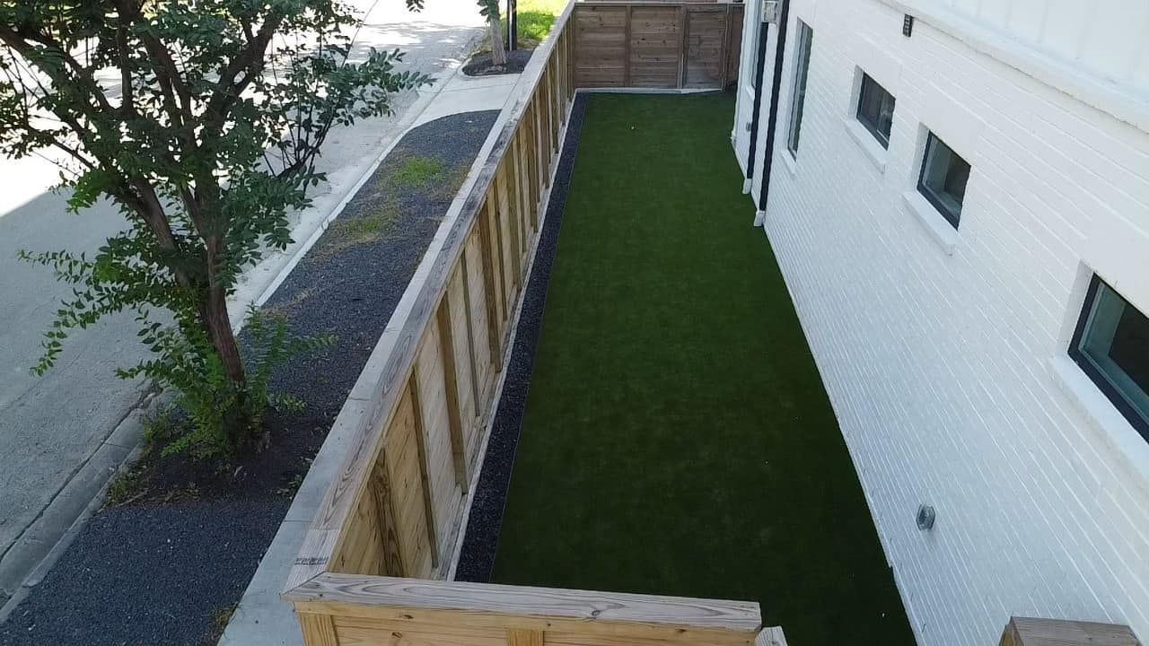 Narrow backyard with artificial turf, wooden fence, and white building with small windows.