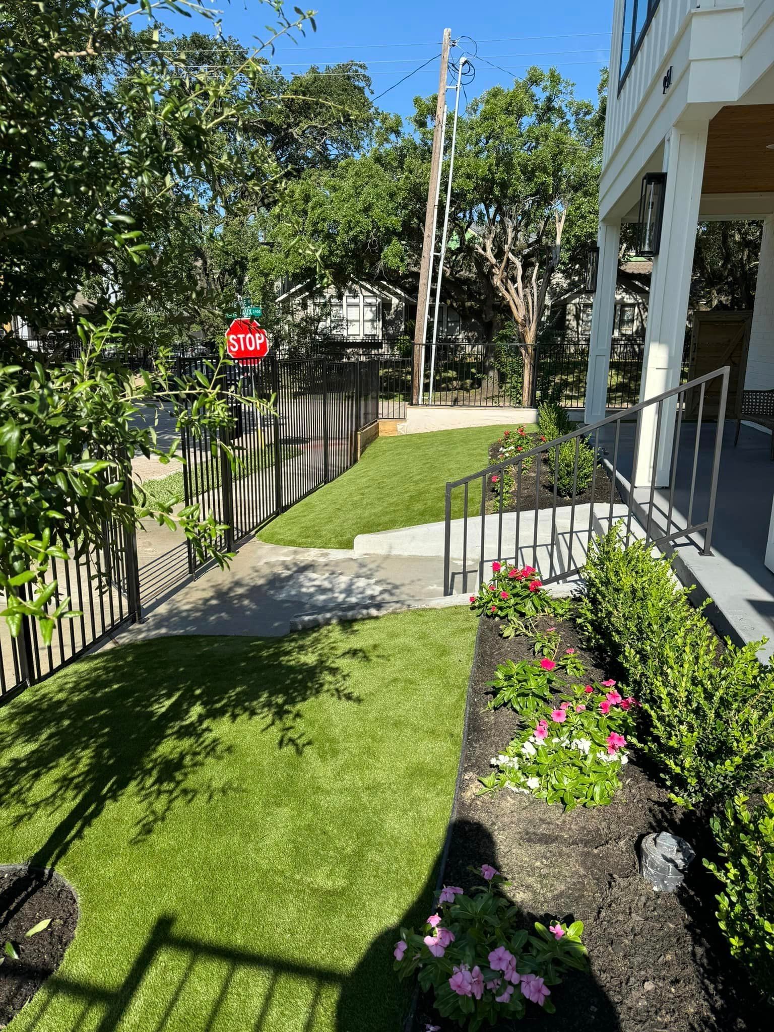 Lawn and garden in front of a house. Black fence, stone path, and flower beds. Bright green grass.