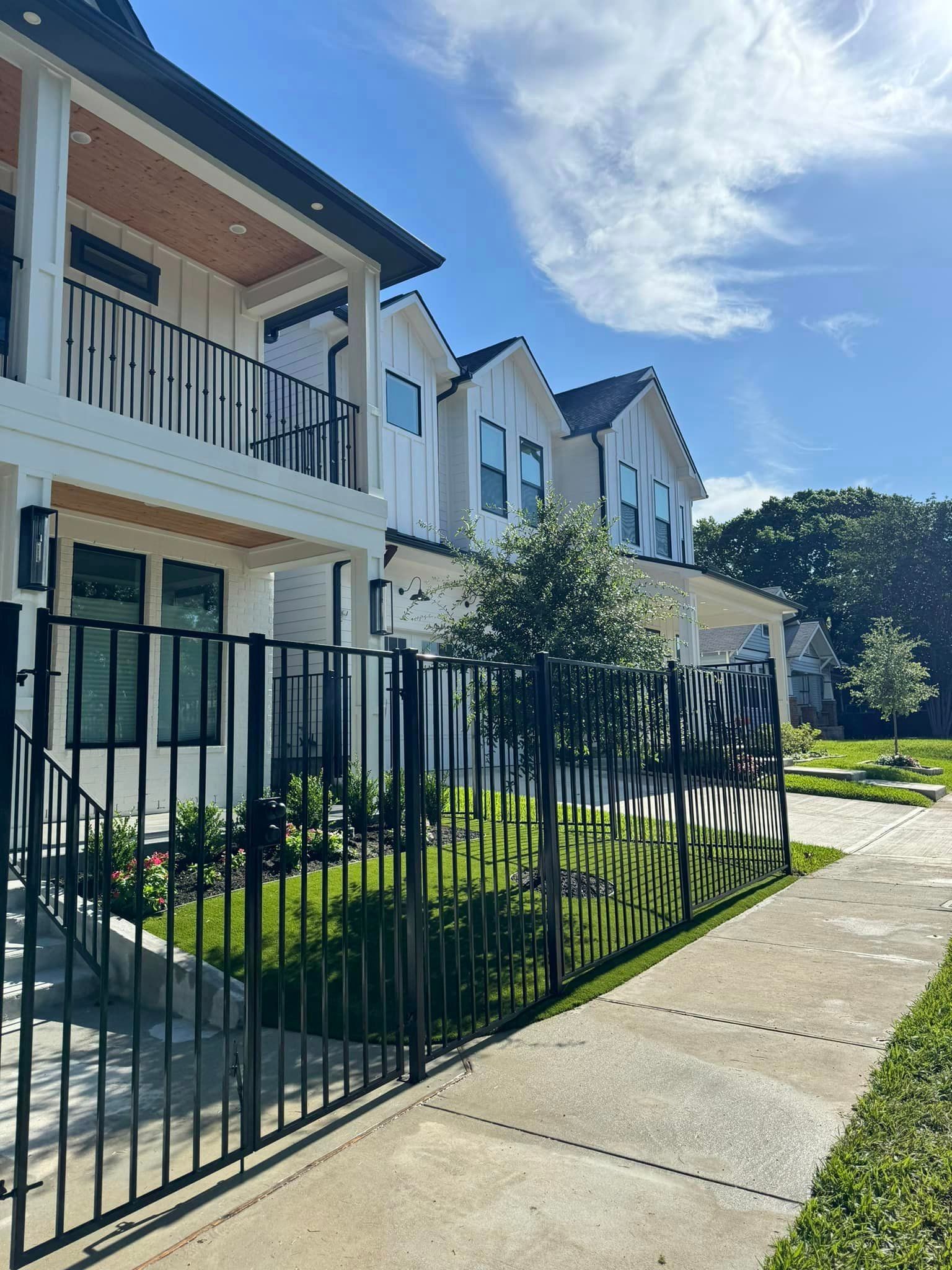 Modern white townhouses behind a black metal fence, sidewalk, green grass, and blue sky.