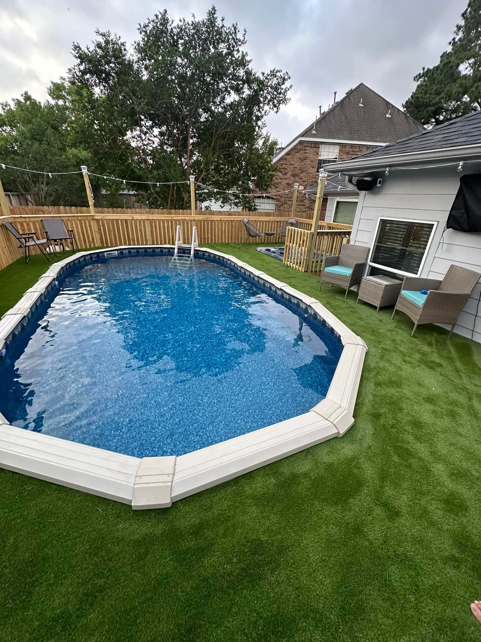Oval above-ground pool with white coping surrounded by green artificial turf. A wooden fence and house are in the background.