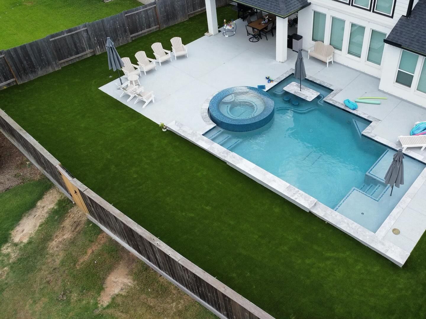 Backyard with pool, jacuzzi, patio, and green lawn. White house in the background, surrounded by a wooden fence.