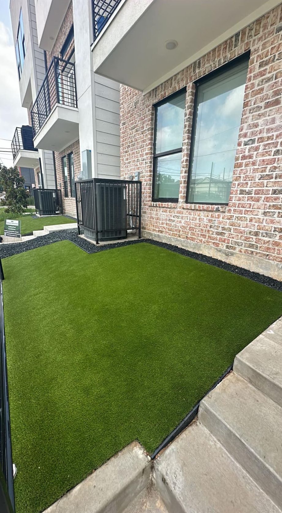 Artificial green lawn next to brick building with an AC unit and a set of stairs.