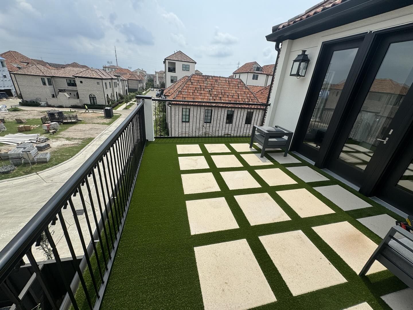 Rooftop patio with green turf and concrete tiles, black railing, and buildings in the background.