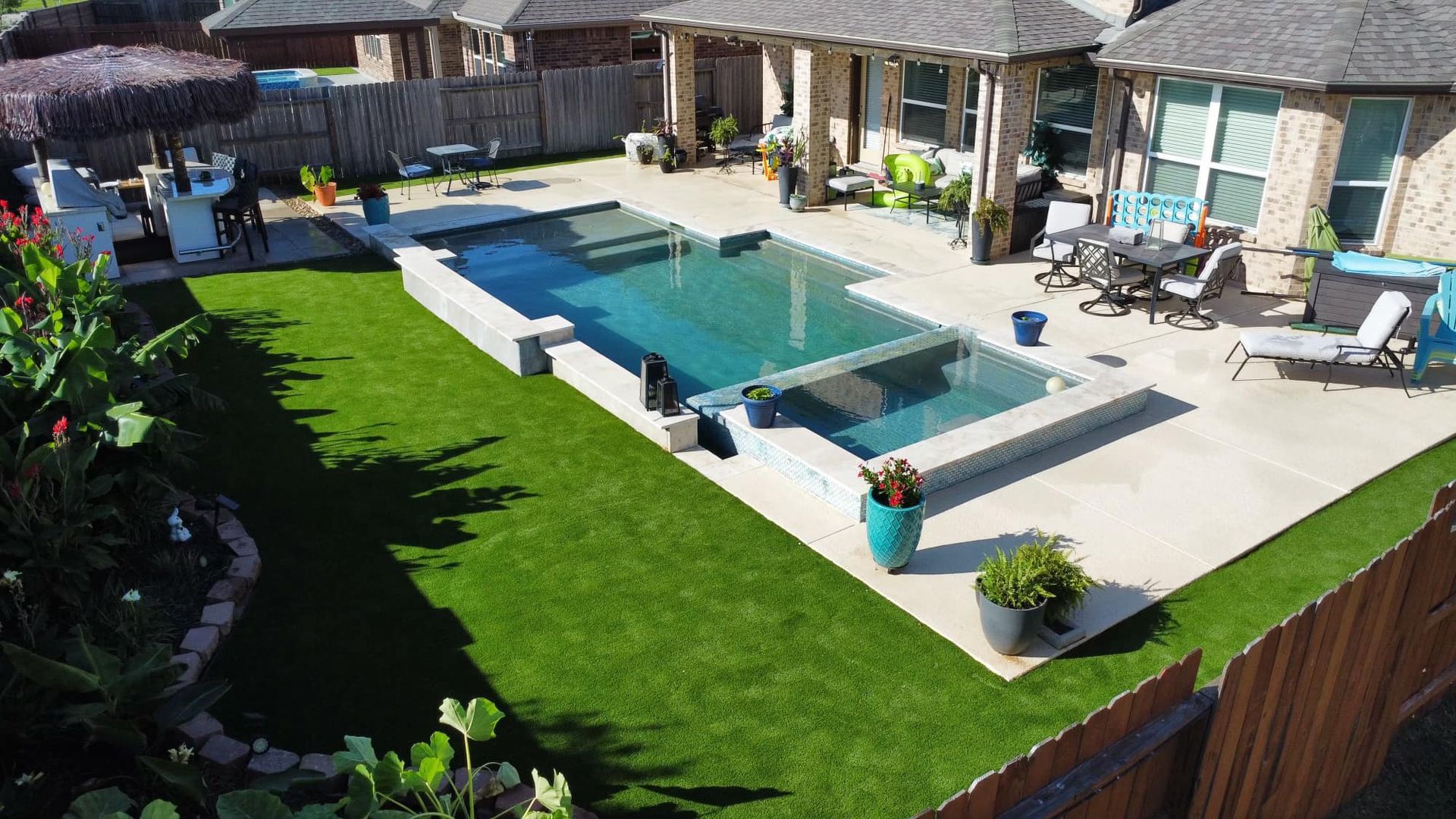Backyard with pool and patio surrounded by green grass. Houses in the background.