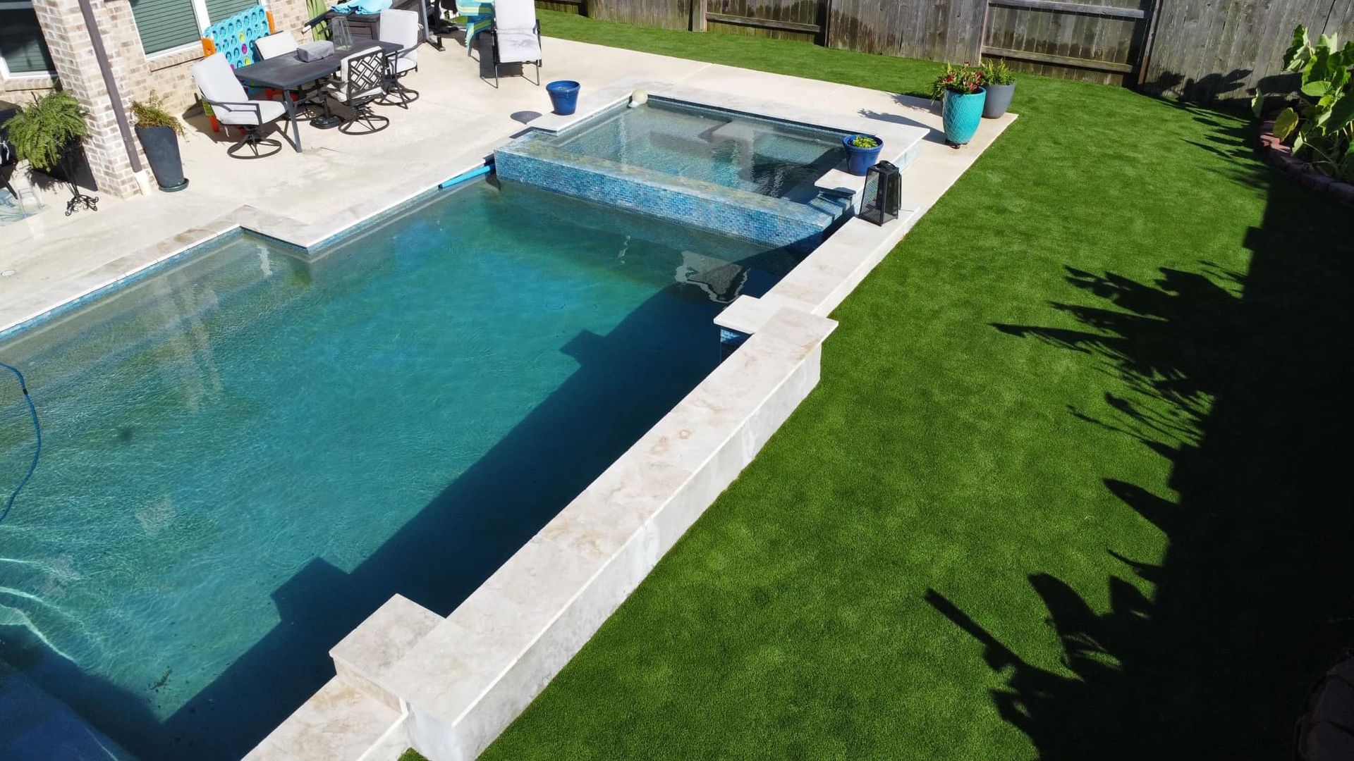 Backyard with a swimming pool, spa, and green lawn. Tables and chairs are near the house.