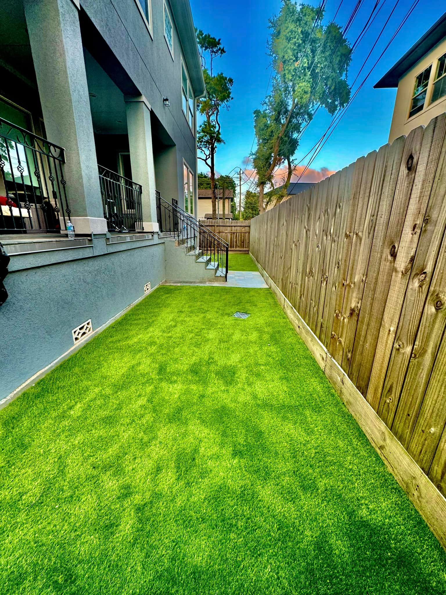 Narrow strip of bright green artificial turf between two buildings, bordered by a wooden fence.