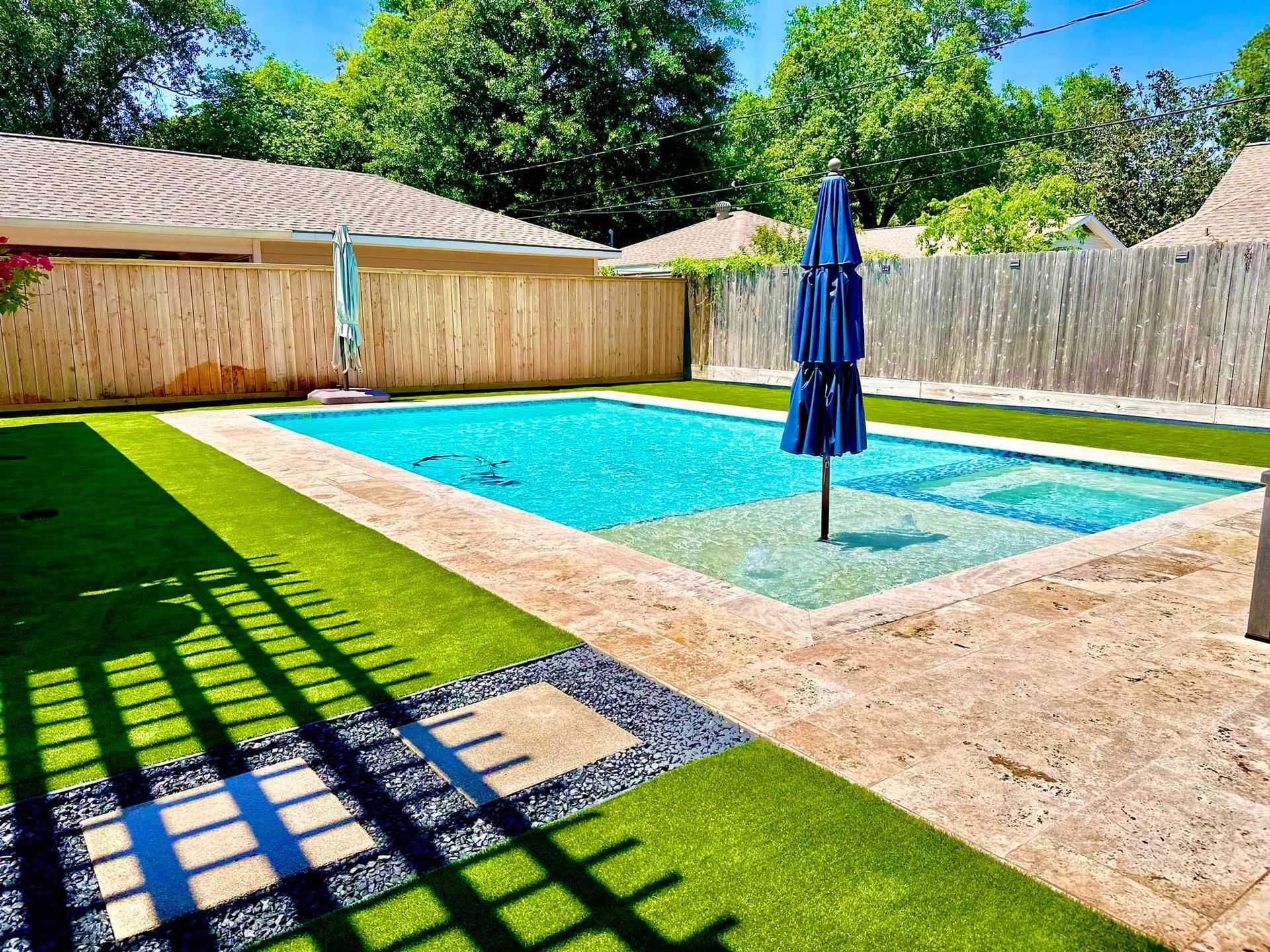Backyard pool with a blue umbrella, surrounded by artificial turf and a wooden fence.