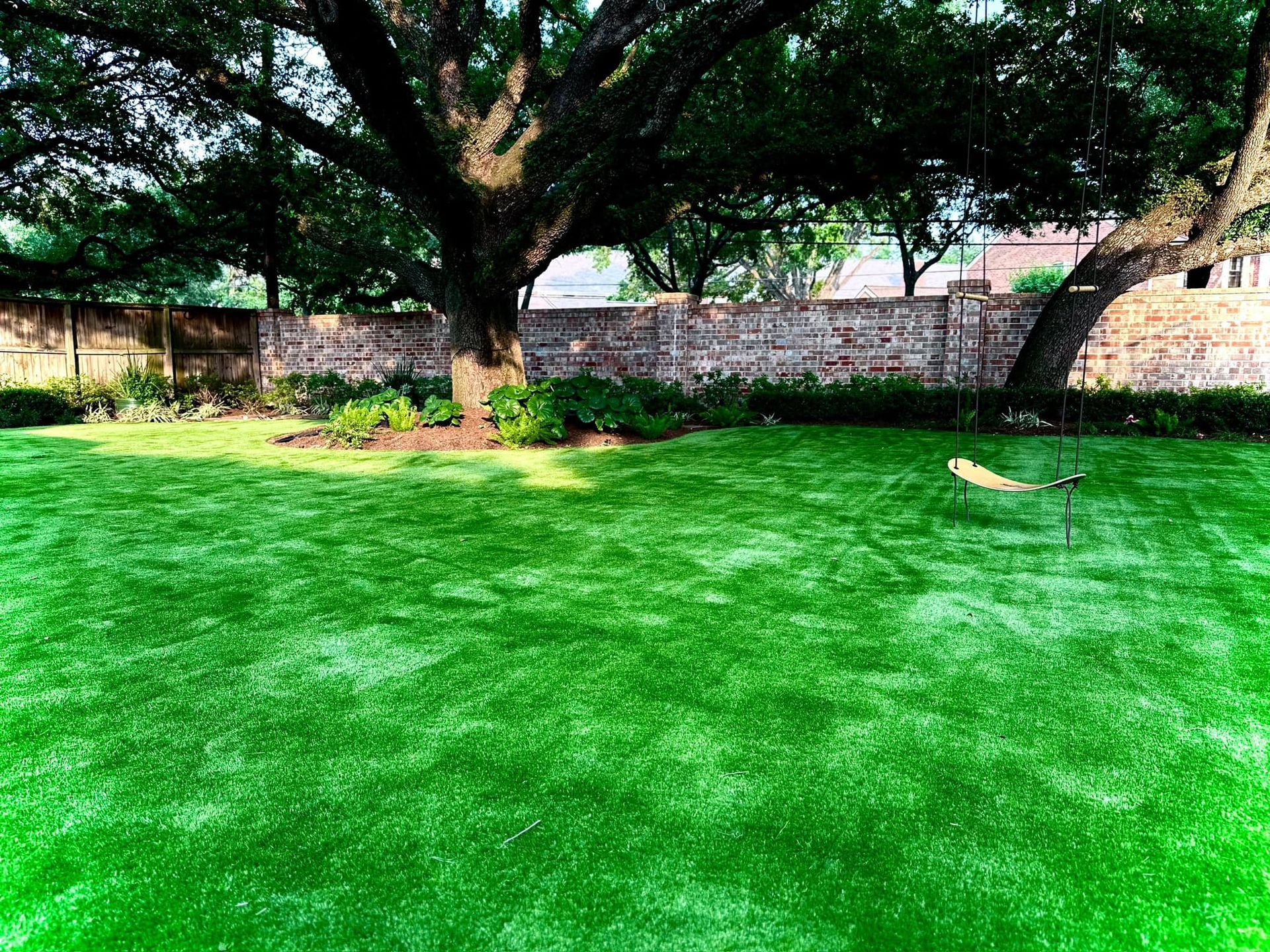Lush green artificial lawn under a large tree with a swing, brick wall in the background.
