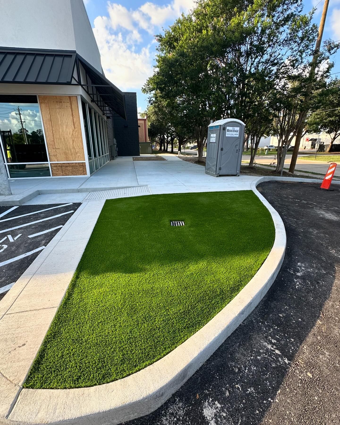 A patch of artificial green grass bordered by concrete. A port-a-potty and building are nearby.