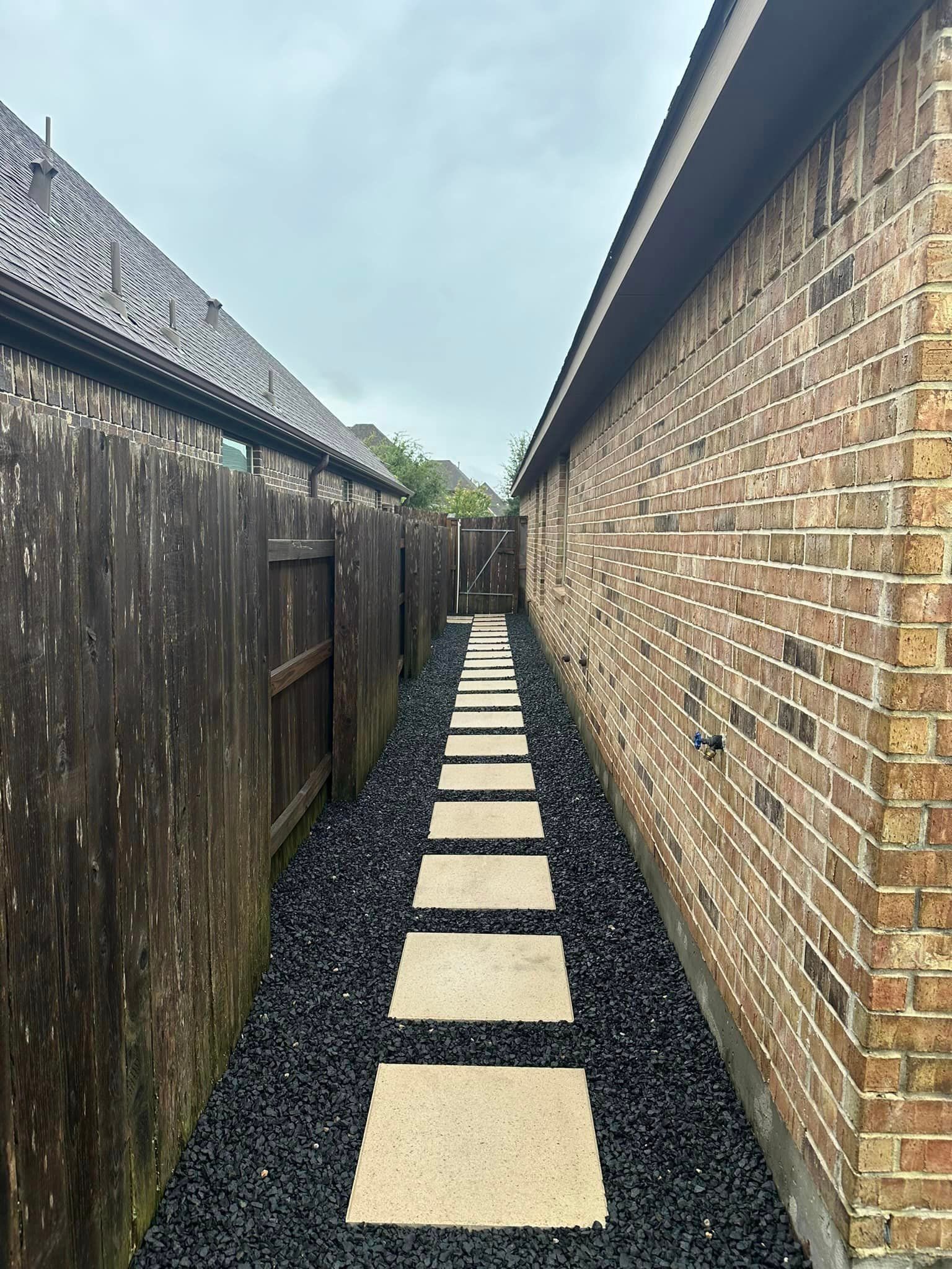 A narrow walkway between a wooden fence and a brick wall. Paved with stepping stones and black rocks.