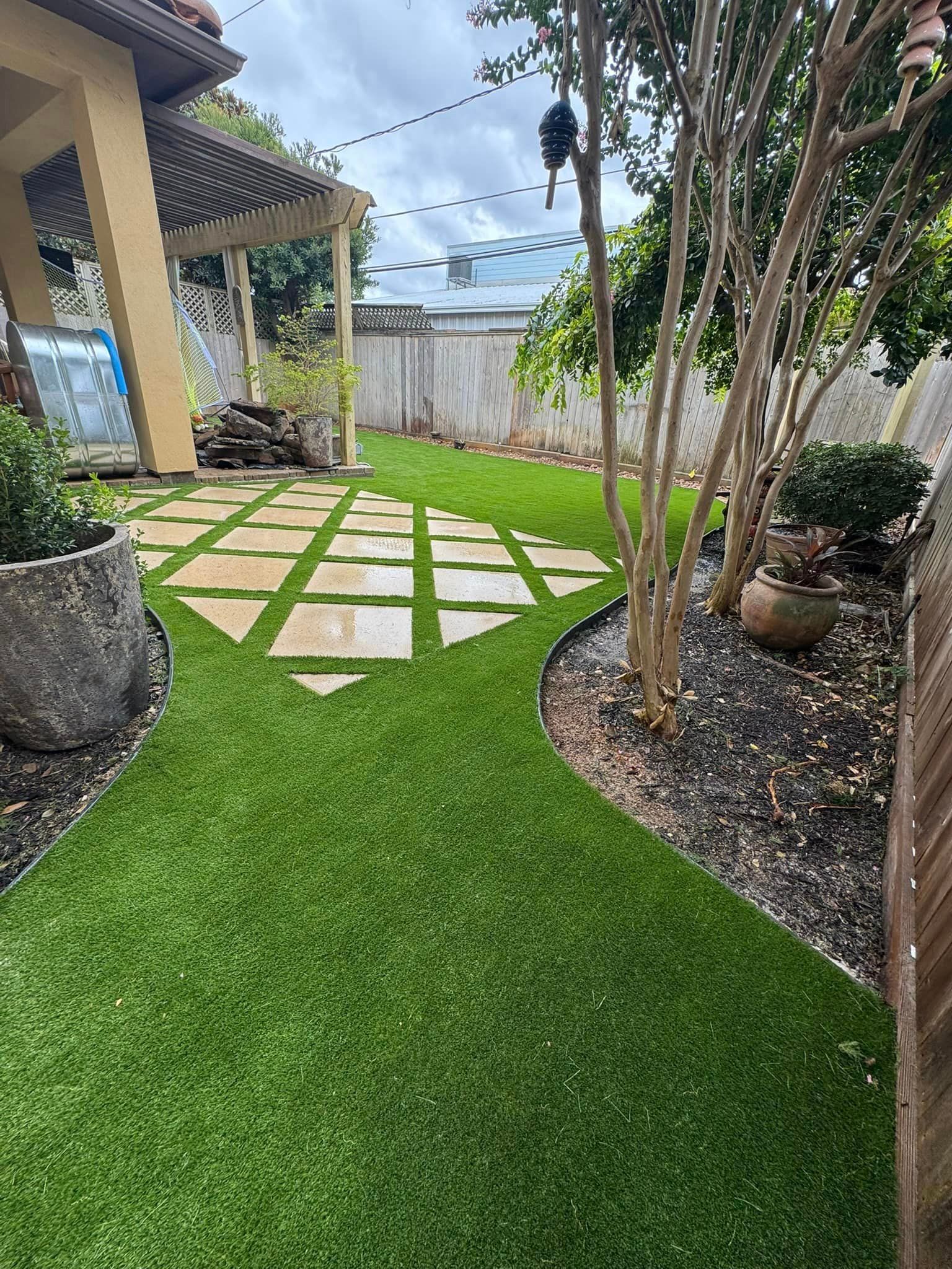 Backyard with a green artificial turf path, stone pavers, and landscaping. A patio and fence are visible.