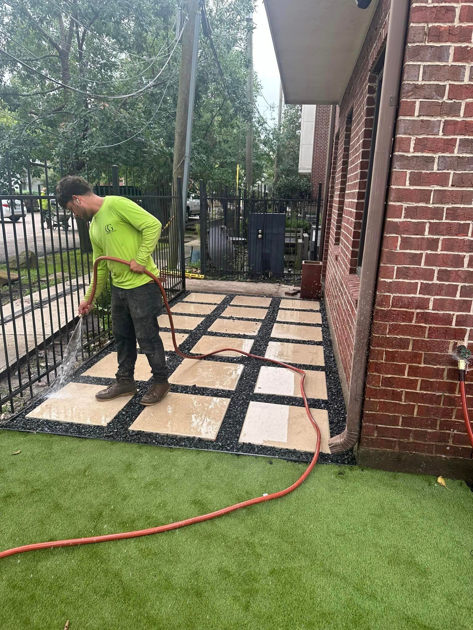 Man watering a stone patio next to a brick building and a black fence.