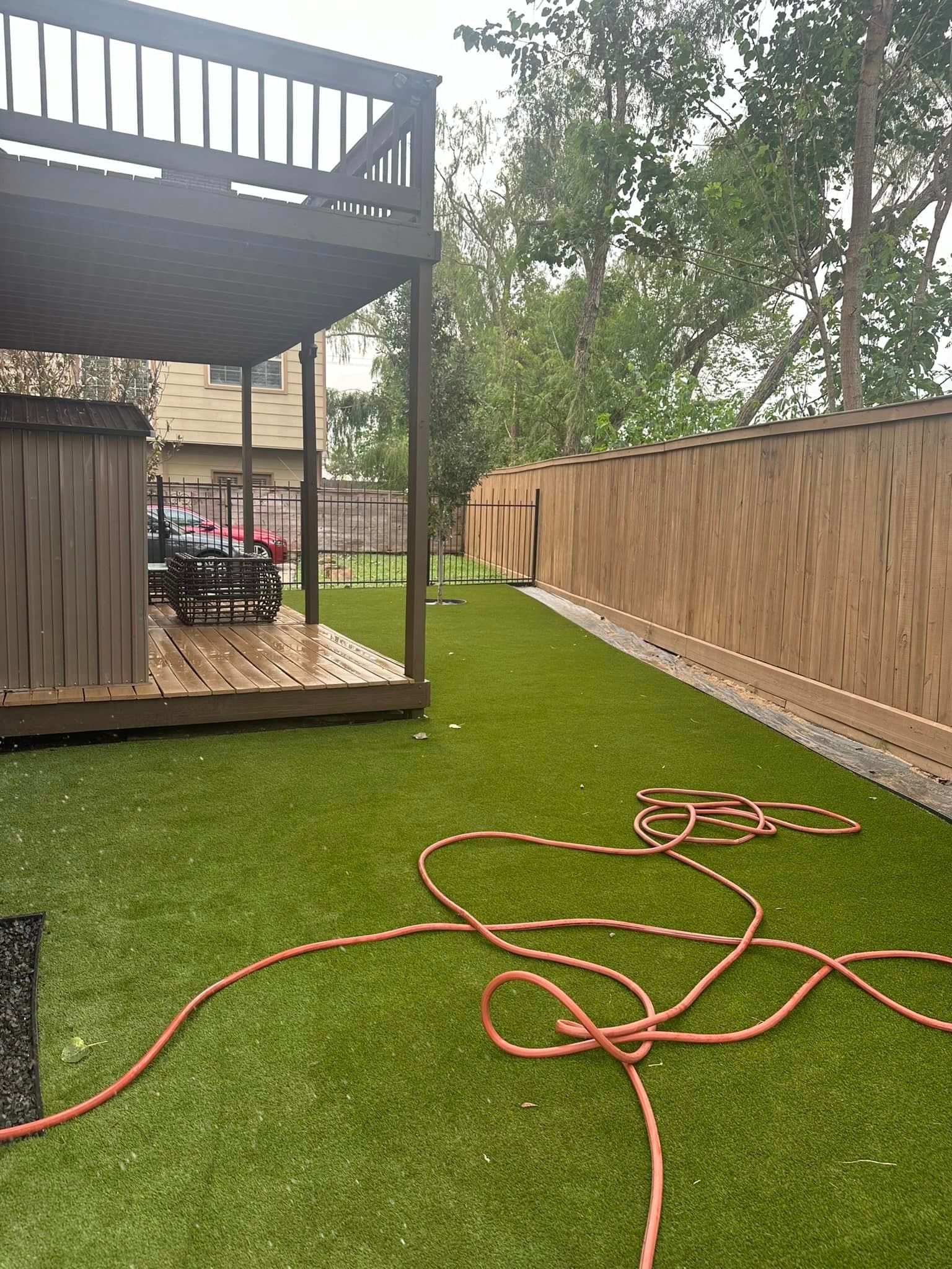 Backyard with artificial turf, wooden deck, brown fence, and orange hose.