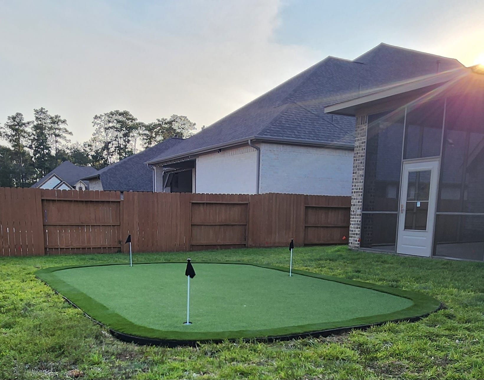 A backyard putting green with three holes and flags, surrounded by grass and a wooden fence.