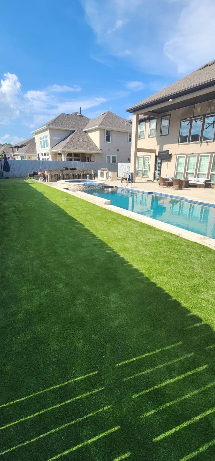 A backyard with green grass, a pool, and houses under a blue sky. Sunlight casts shadows on the lawn.