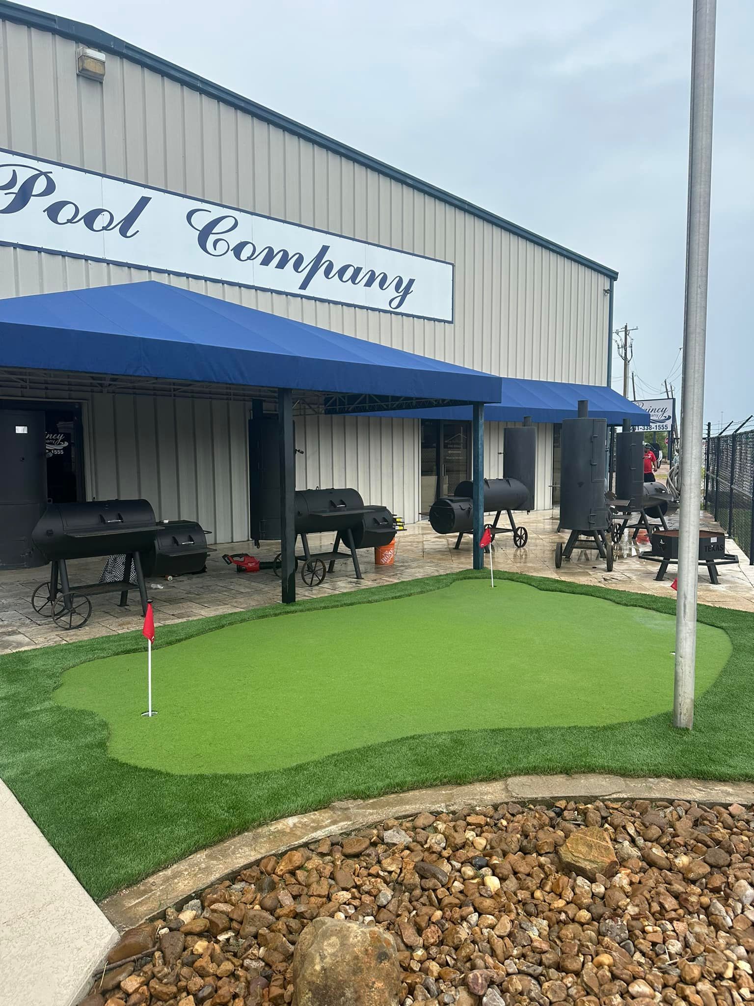 Exterior of J Pool Company with BBQs under blue awnings, a putting green, and a building with a sign.