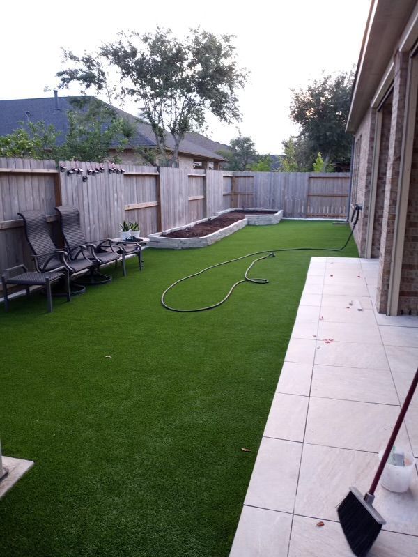 Green artificial turf backyard with patio, fence, chairs, and garden beds.