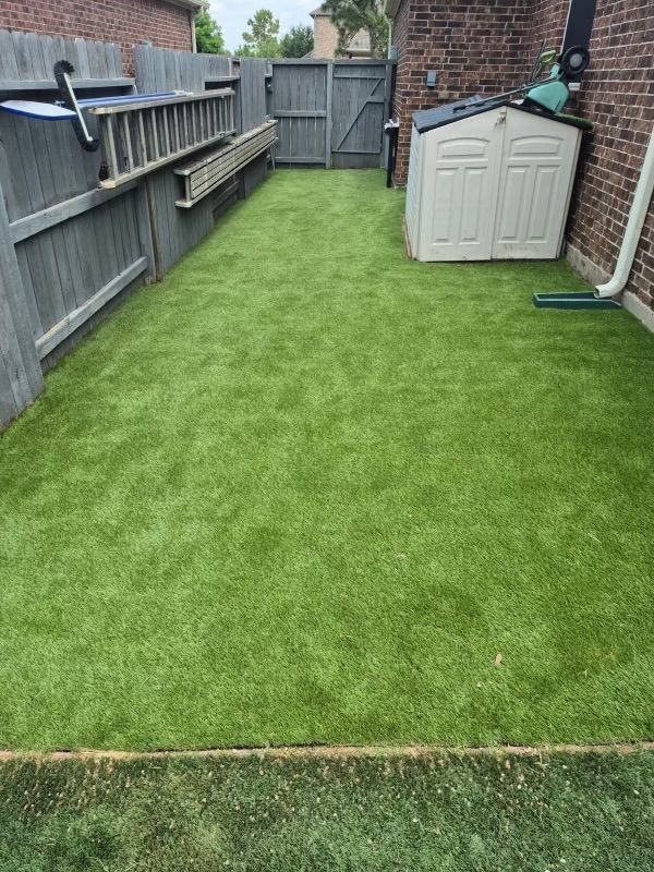 Narrow backyard with artificial turf, bordered by a wooden fence, a shed, and a brick wall.