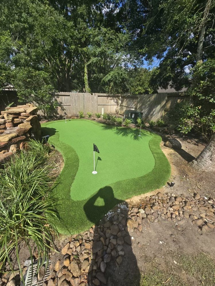 Backyard putting green with a black flag and green turf, surrounded by rocks and grass.
