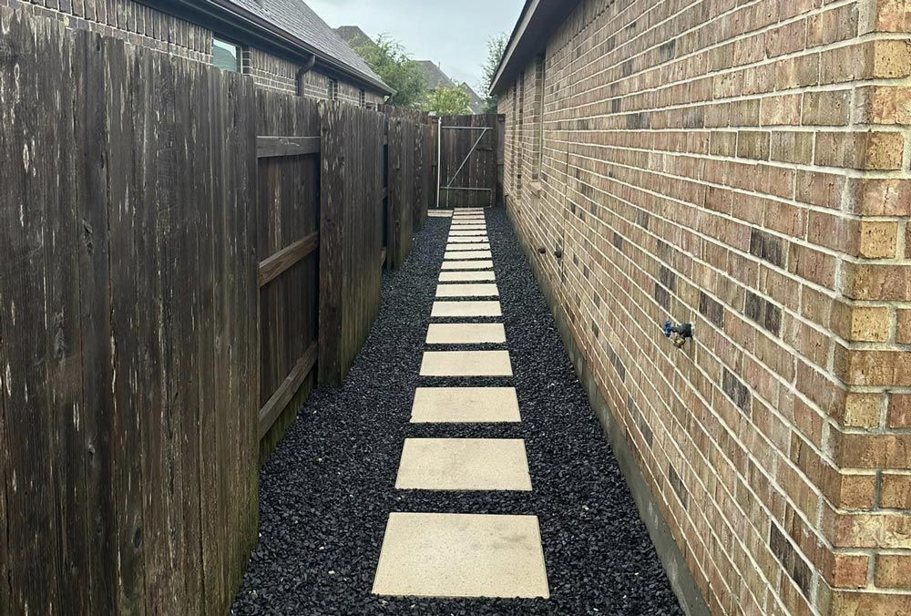 Narrow walkway between a brick building and a wooden fence. Stepping stones lead to a gate. Dark gravel.