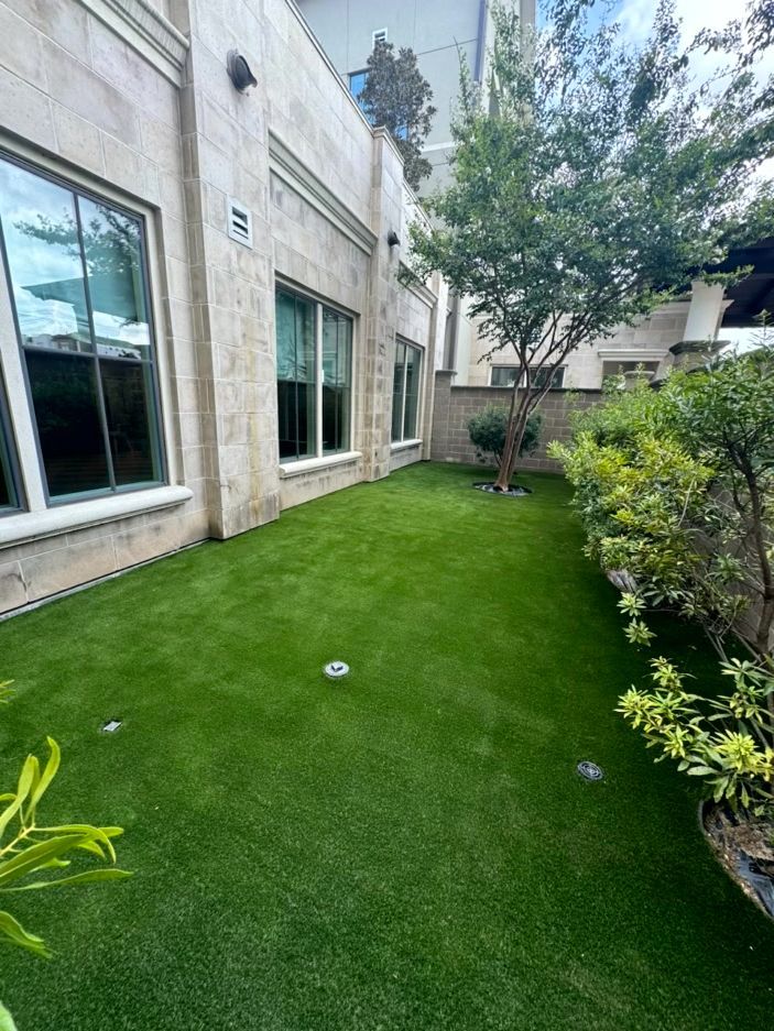 Green artificial turf in a small yard between a building with windows and bushes, with a tree in the background.