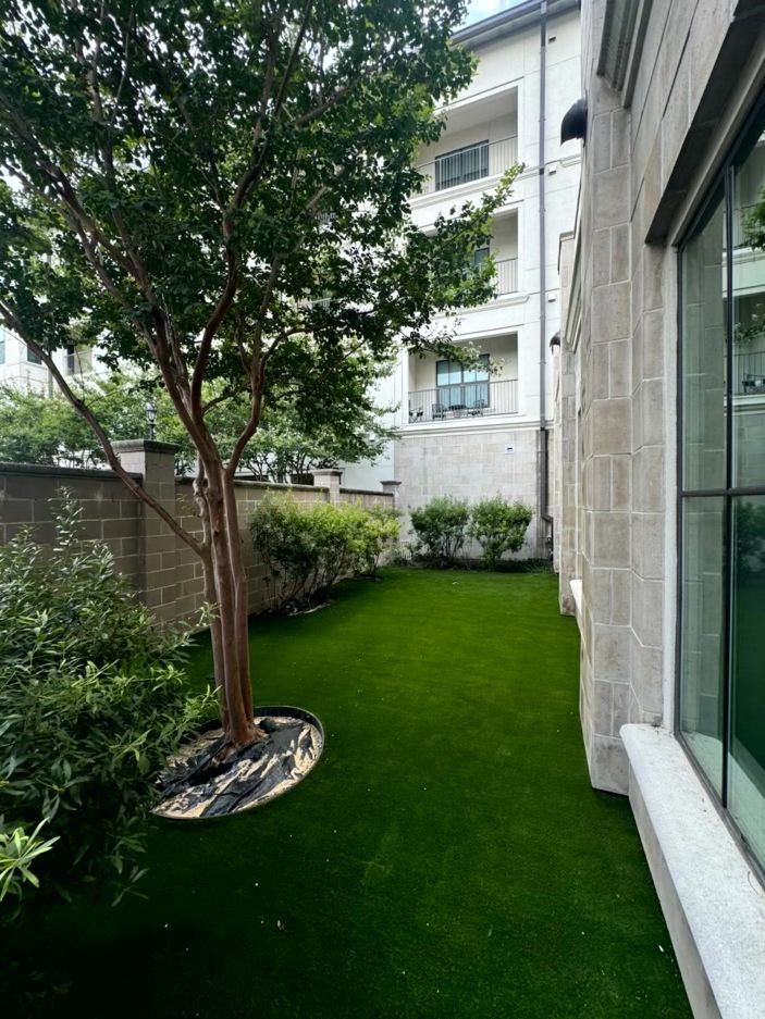 Lush green artificial grass in a narrow courtyard between a stone building and a brick wall, with a tree and bushes.